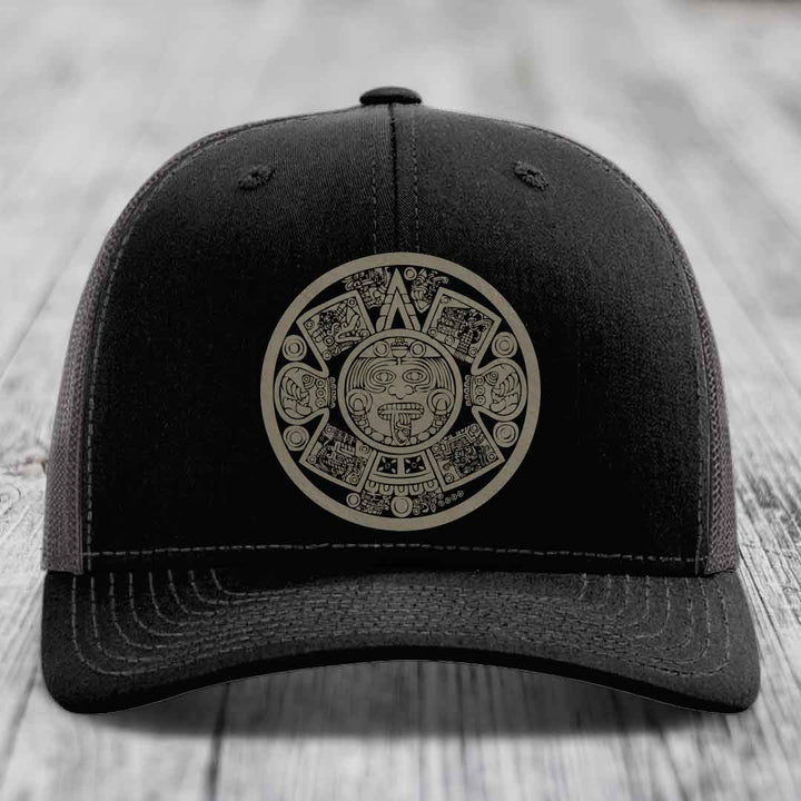 Black trucker hat with Aztec calendar design on front, displayed on wooden surface.