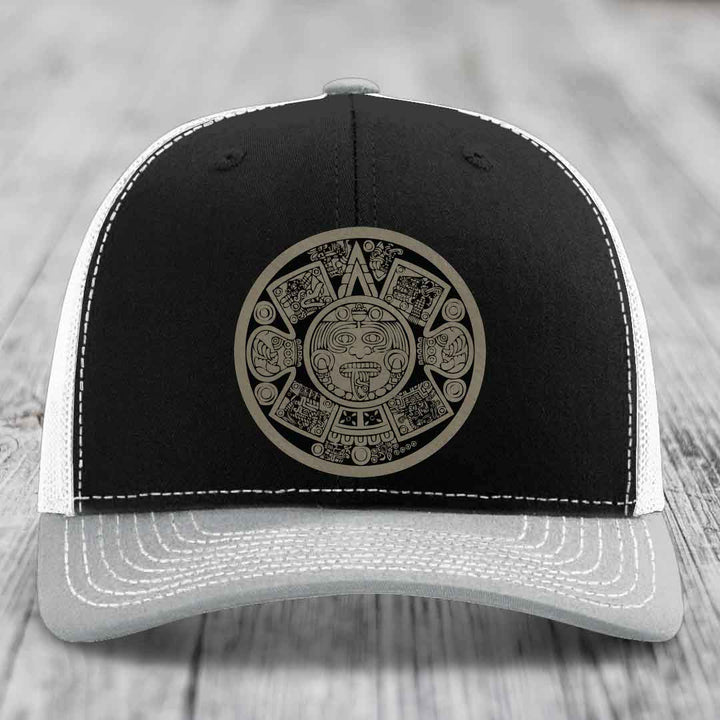 Black and grey trucker hat with Aztec calendar patch on front.