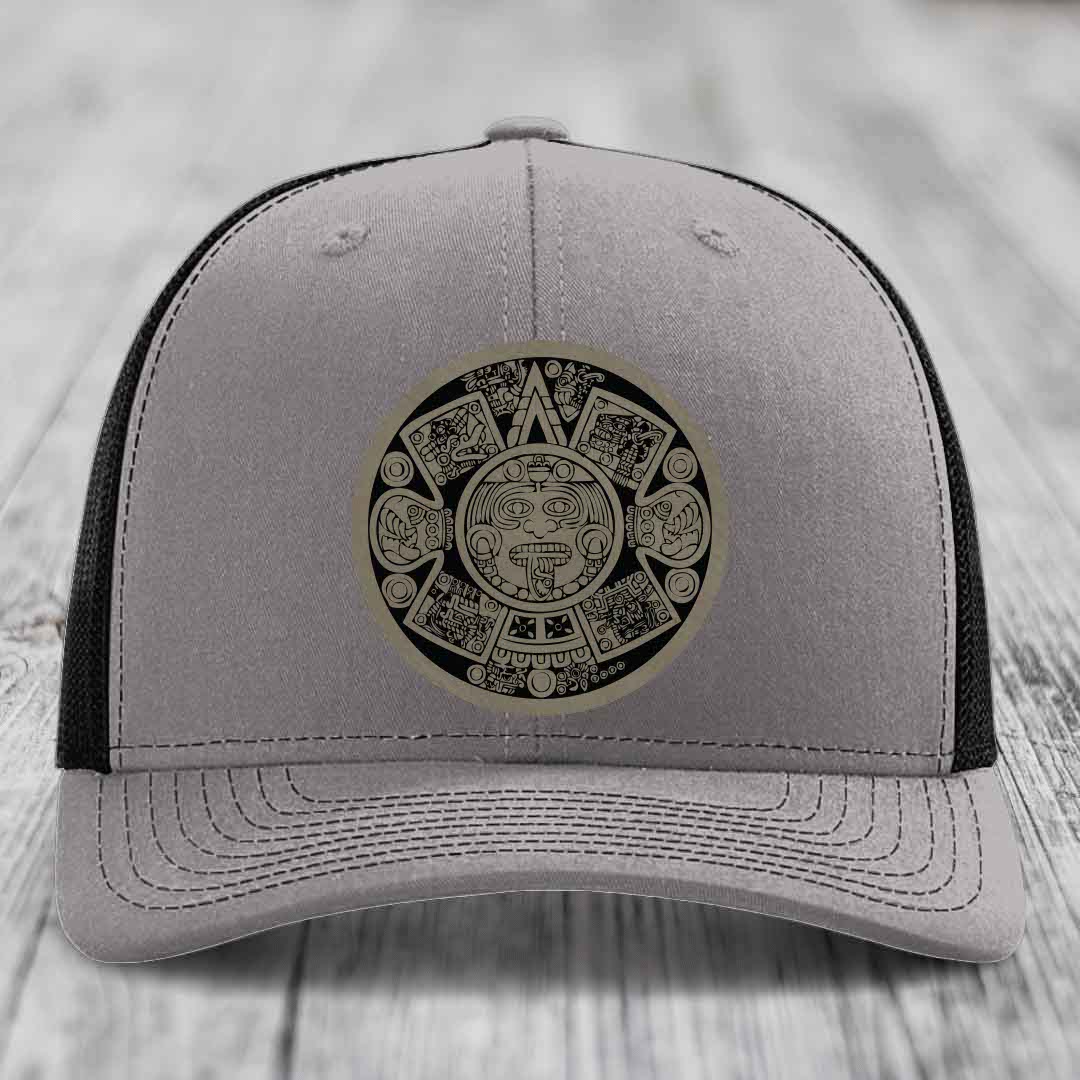 Grey trucker hat with black mesh back and an Aztec calendar leather patch on the front.