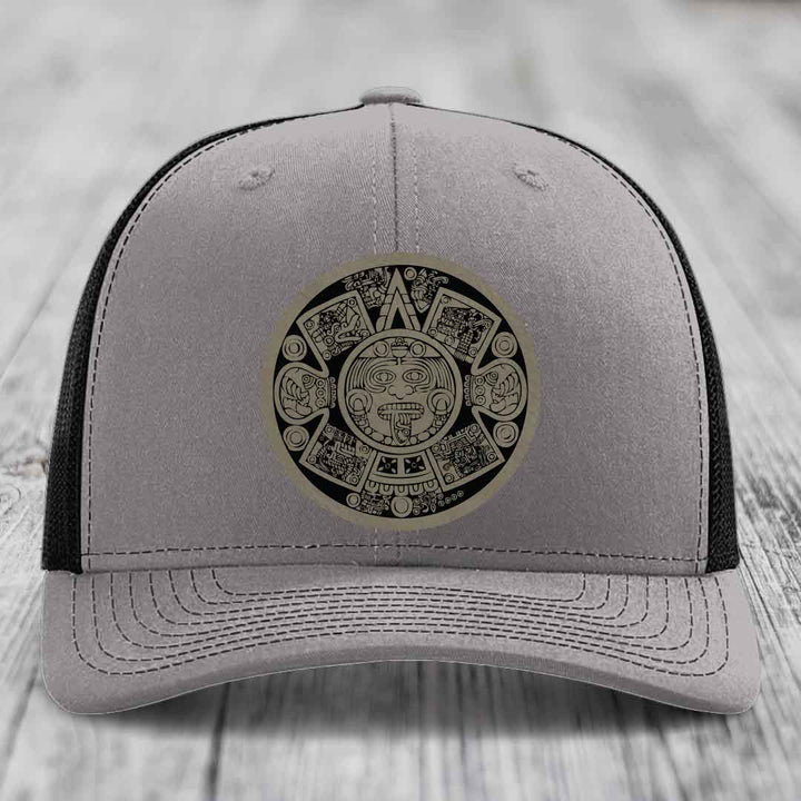 Grey trucker hat with black mesh back and an Aztec calendar leather patch on the front.