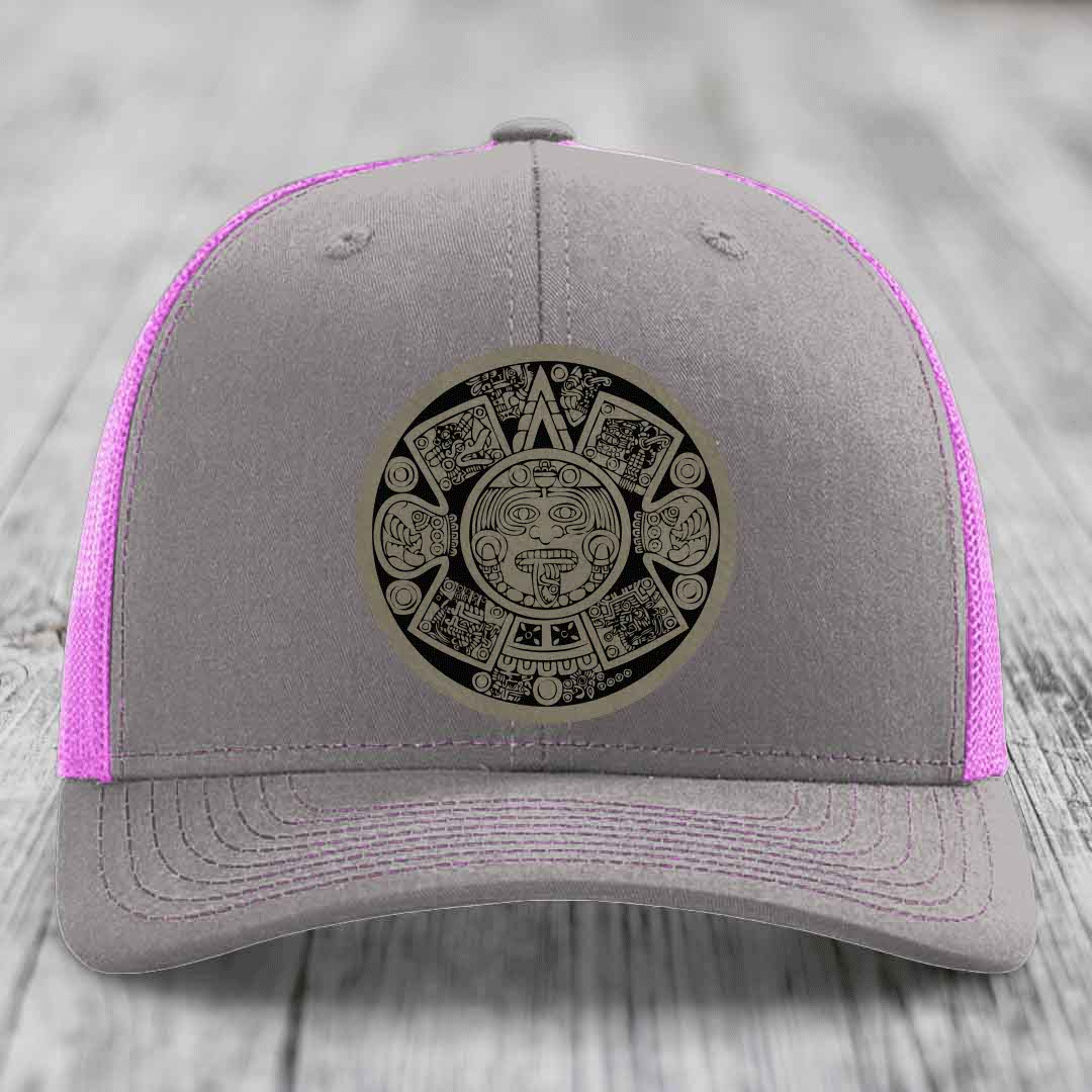 Grey trucker hat with pink mesh and stitching, featuring a black Aztec calendar patch on front.