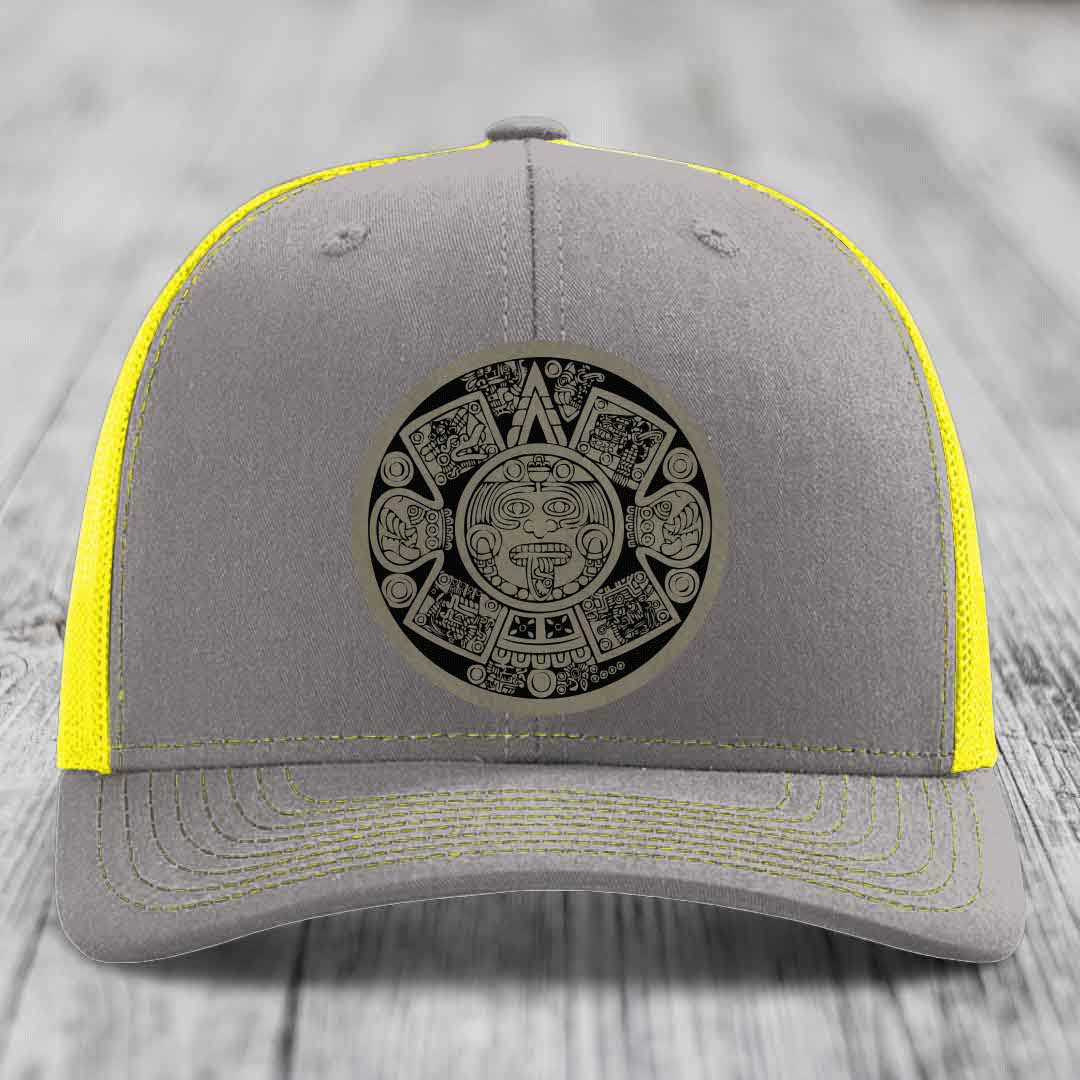 Grey trucker hat with yellow mesh and stitching, featuring a black Aztec calendar patch on front.