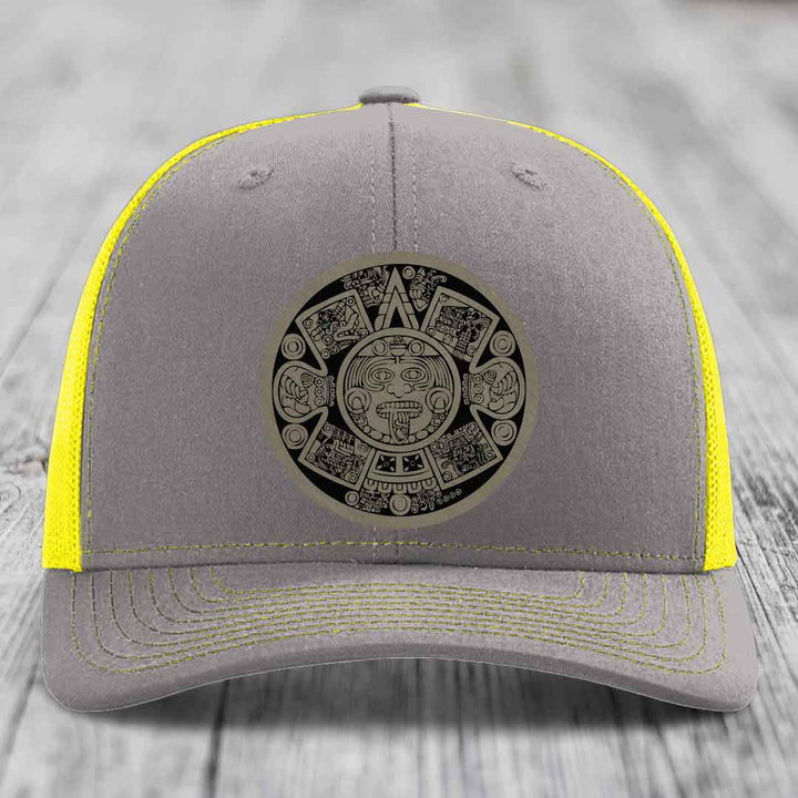 Grey trucker hat with yellow mesh and stitching, featuring a black Aztec calendar patch on front.