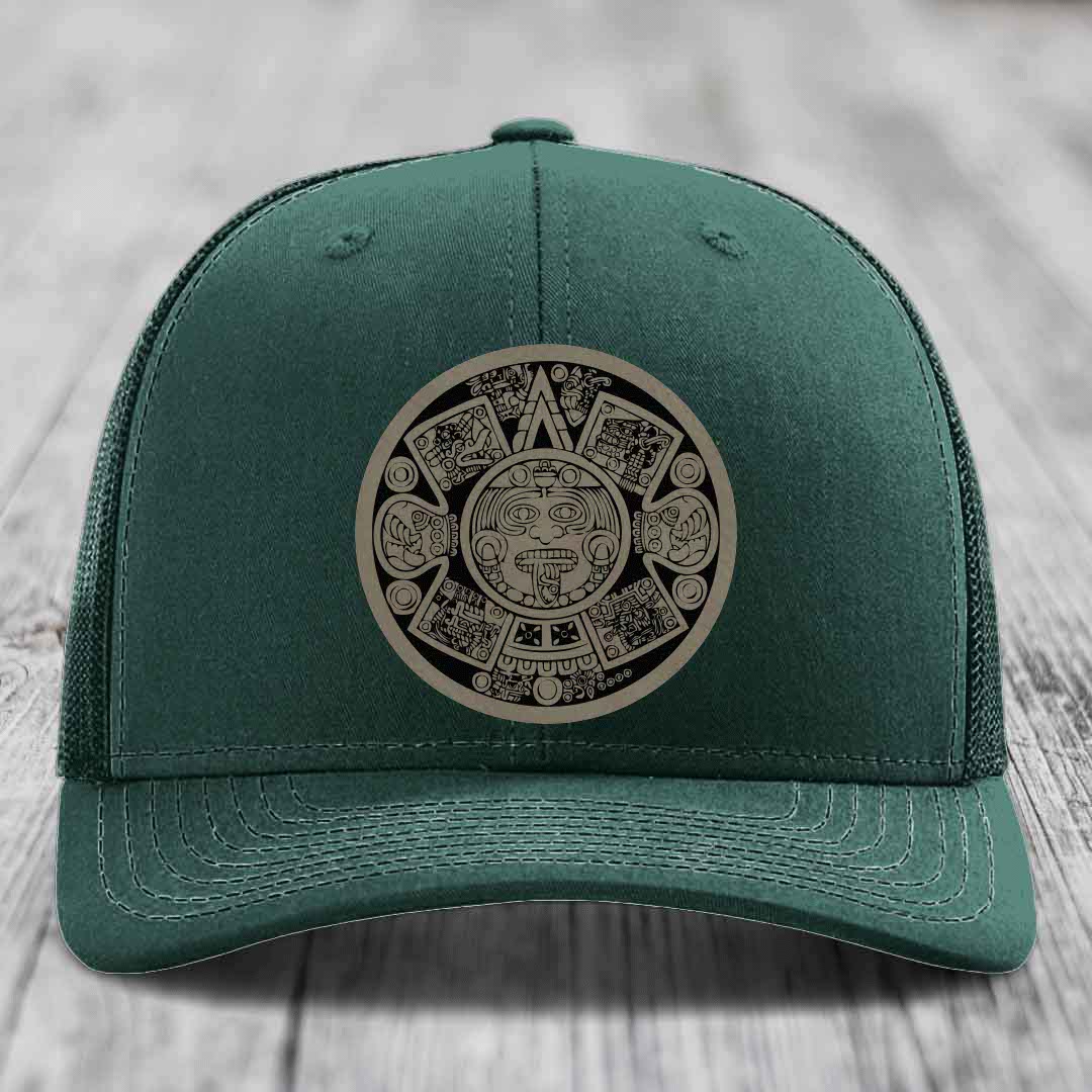 Green trucker hat with Aztec calendar patch on front, displayed on wooden surface.