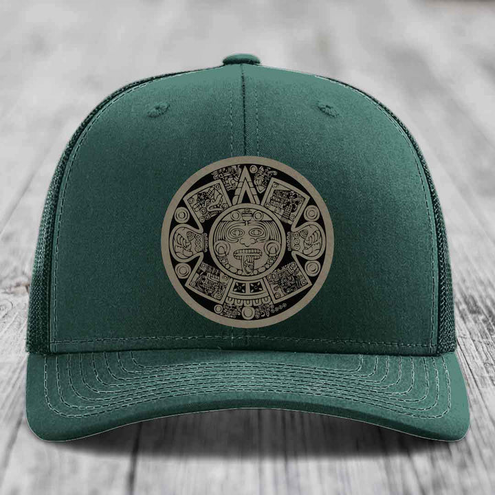 Green trucker hat with Aztec calendar patch on front, displayed on wooden surface.