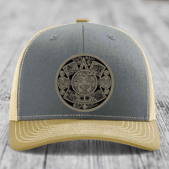 Grey and beige trucker hat with Aztec calendar patch on front.