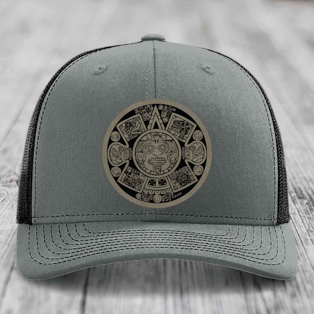 Grey trucker hat with Aztec calendar leather patch on front, black mesh back.