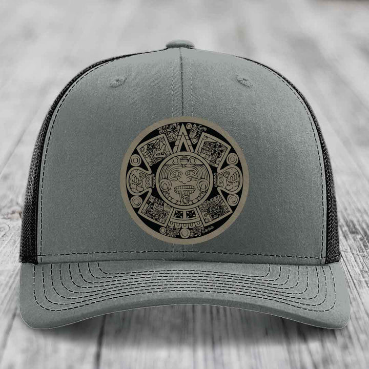 Grey trucker hat with Aztec calendar leather patch on front, black mesh back.