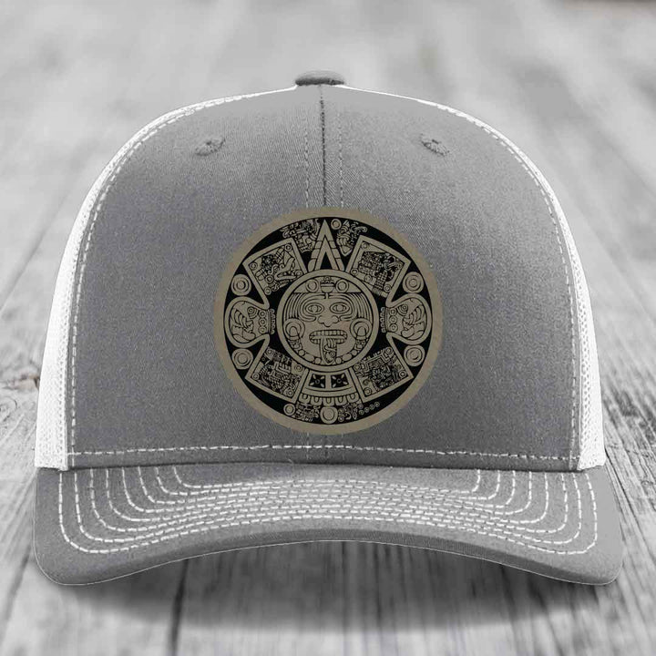 Grey trucker hat with white mesh back and stitching, featuring a circular Aztec calendar leather patch.