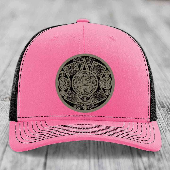 Pink trucker hat with black mesh back and gray Aztec calendar patch on front.