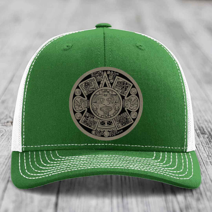 Green trucker hat with white mesh back and stitching, featuring an Aztec calendar leather patch.