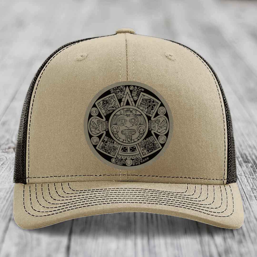 Beige trucker hat with black Aztec calendar patch on front, black mesh back, and curved brim.