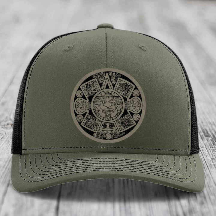 Olive green trucker hat with Aztec calendar patch on front, black mesh back.