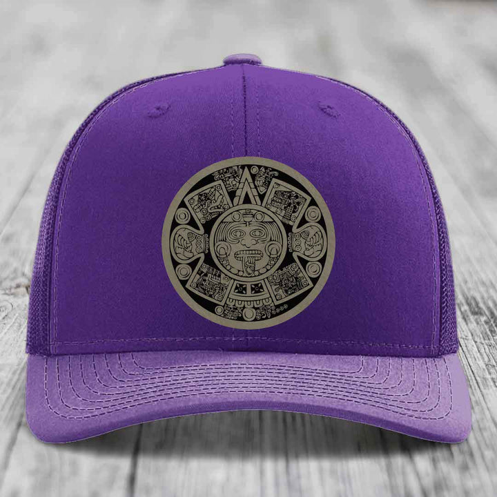 Purple trucker hat with Aztec calendar patch on front, displayed on wooden surface.
