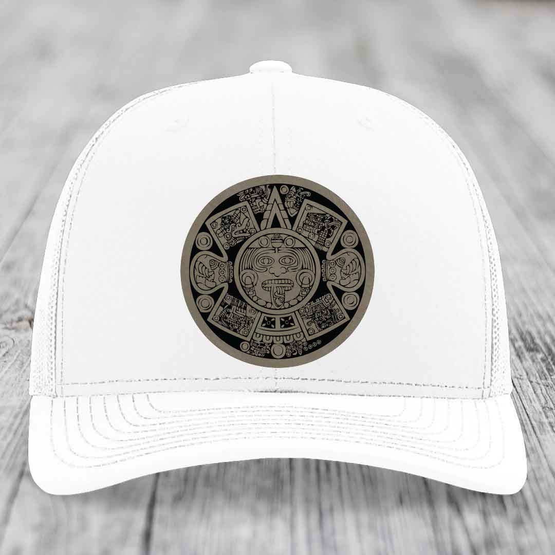 White trucker hat with Aztec calendar patch on front, resting on wooden surface.