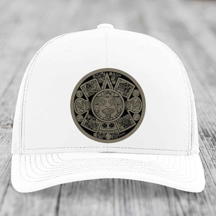 White trucker hat with Aztec calendar patch on front, resting on wooden surface.