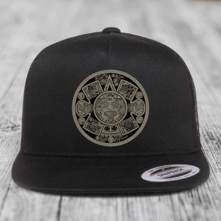 Black snapback hat with Aztec calendar design patch on front, displayed on wooden surface.