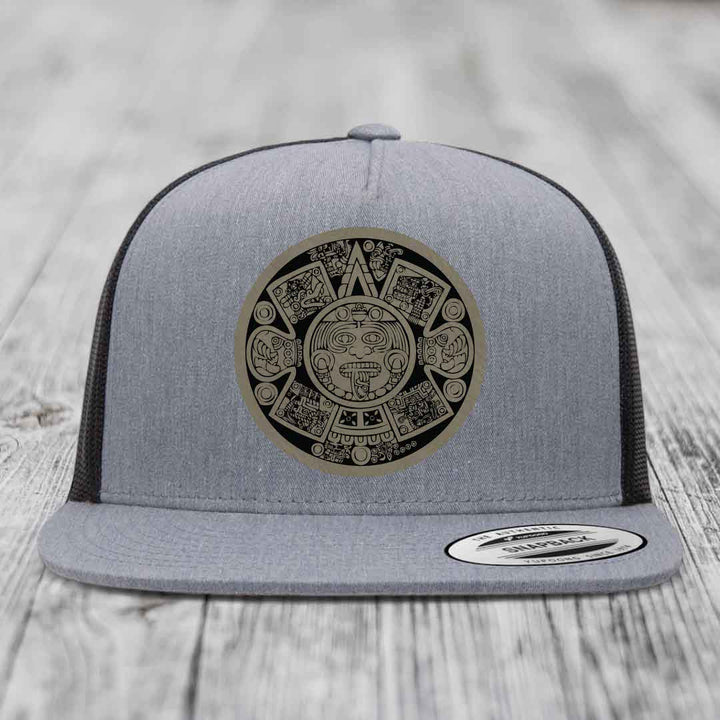 Grey trucker hat with Aztec calendar patch on front, black mesh back, flat brim.