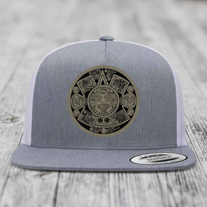 Grey trucker hat with Aztec calendar patch on front, white mesh back, flat brim.