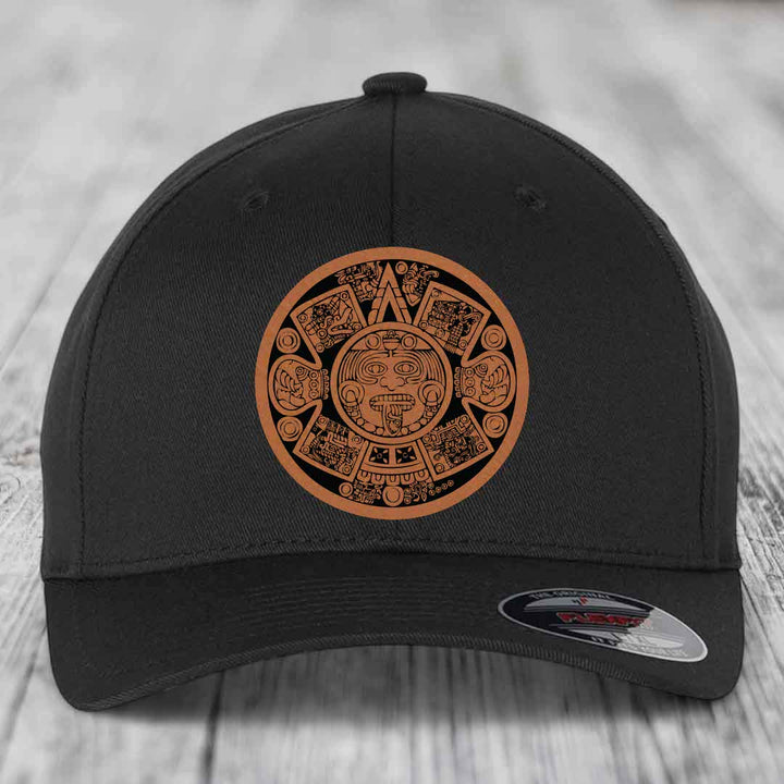 Black baseball cap with brown Aztec calendar patch on front, displayed on wooden surface.