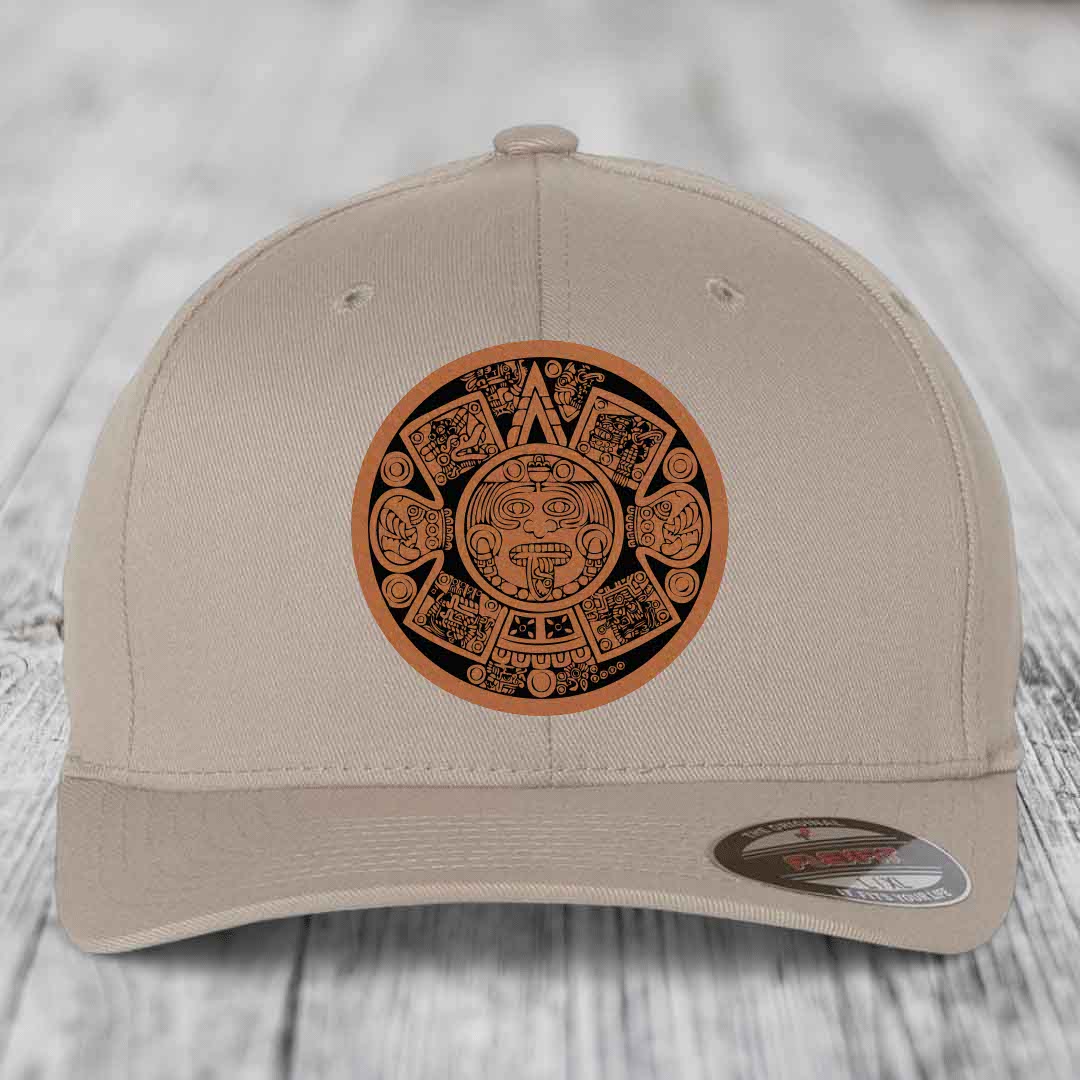 Beige baseball cap with Aztec calendar leather patch on front, displayed on wooden surface.
