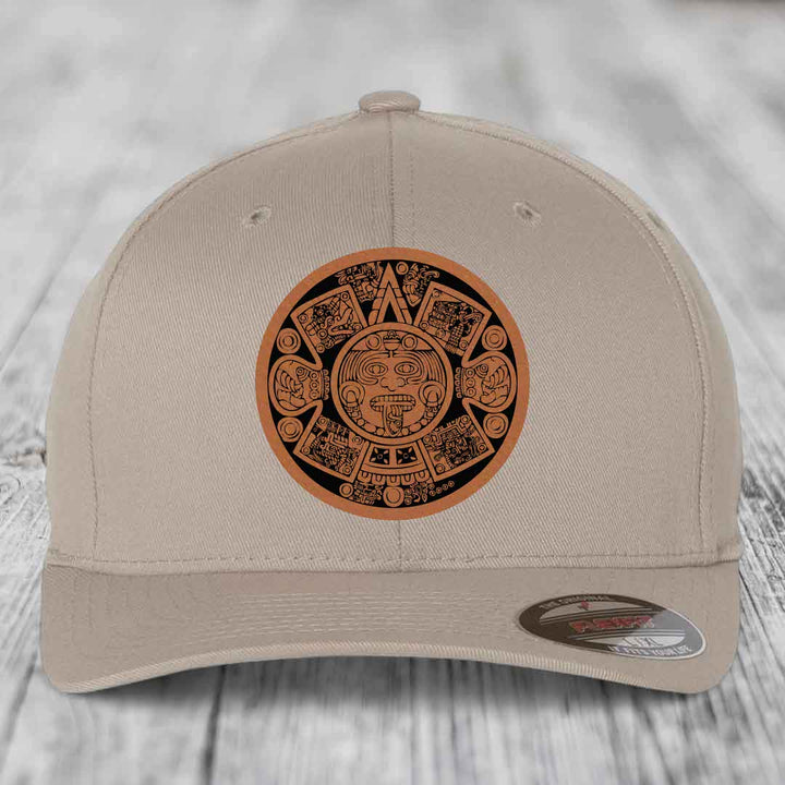 Beige baseball cap with Aztec calendar leather patch on front, displayed on wooden surface.