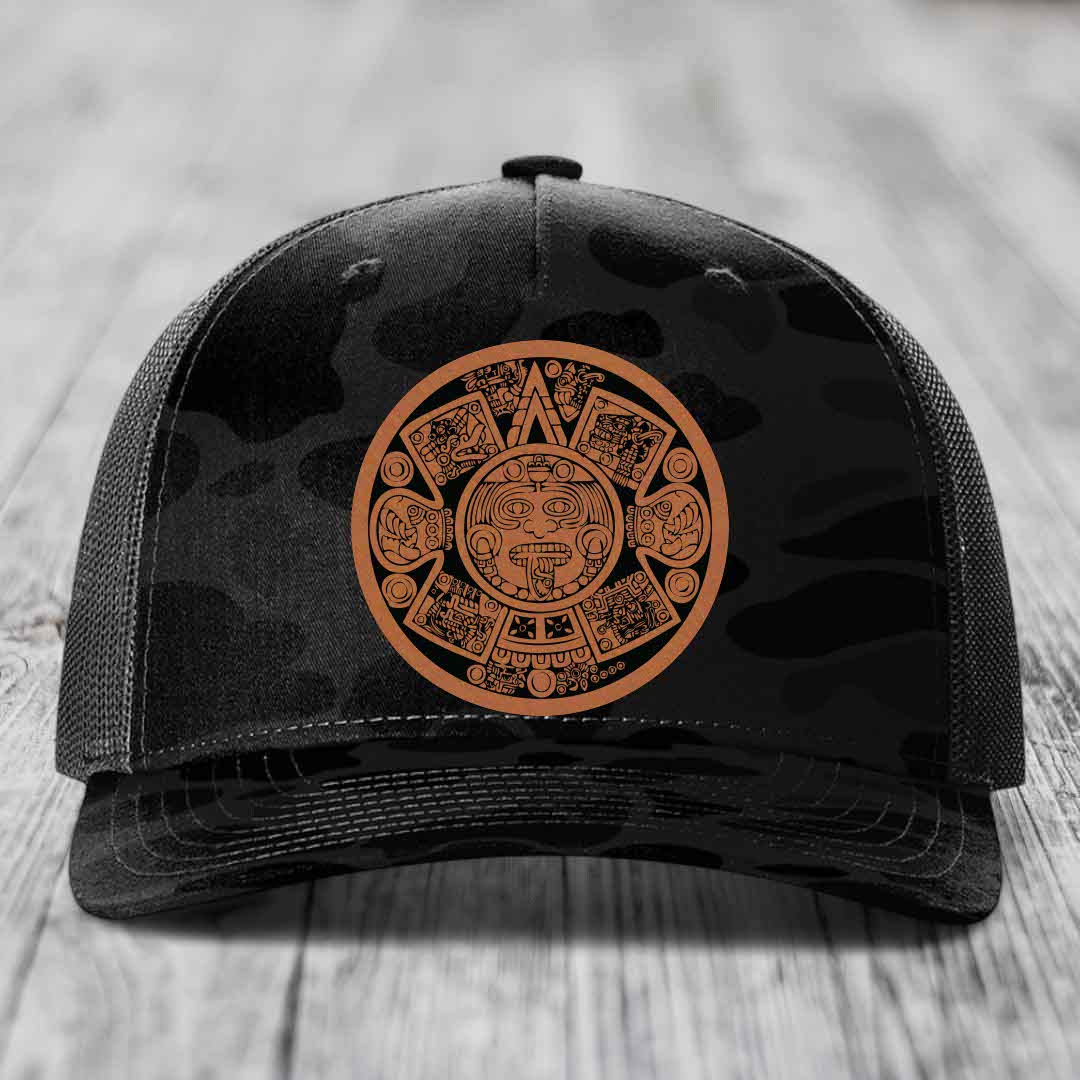 Black trucker hat with brown Aztec calendar patch on front, resting on wooden surface.