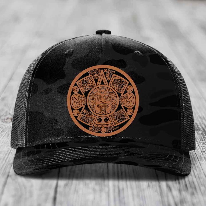 Black trucker hat with brown Aztec calendar patch on front, resting on wooden surface.