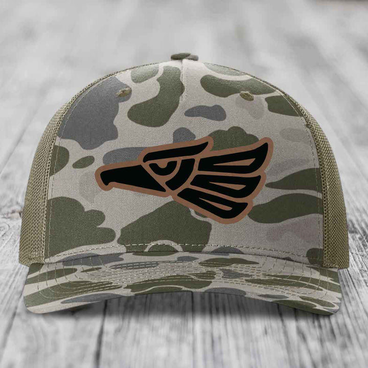 Camouflage trucker hat with black and brown Aztec eagle patch on front.