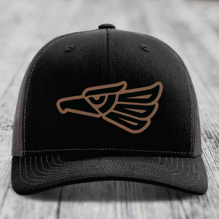 Black trucker hat with brown Aztec-style eagle patch on front, resting on wooden surface.