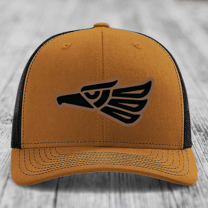 Tan trucker hat with black Aztec-style eagle patch on front, black mesh back and adjustable strap.
