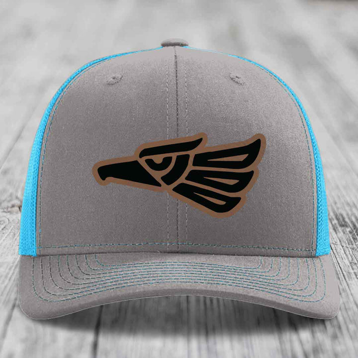 Grey trucker hat with blue mesh sides and a black Aztec-style eagle patch.
