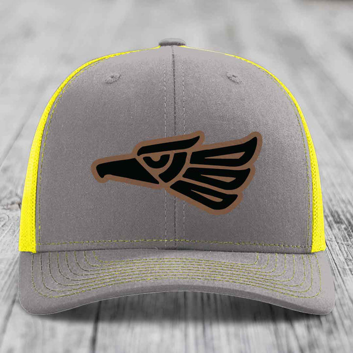 Grey trucker hat with yellow mesh and stitching, featuring a black and brown Aztec-style eagle patch.