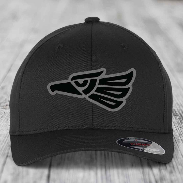 Black baseball cap with Aztec-style eagle patch on front, displayed on wooden surface.