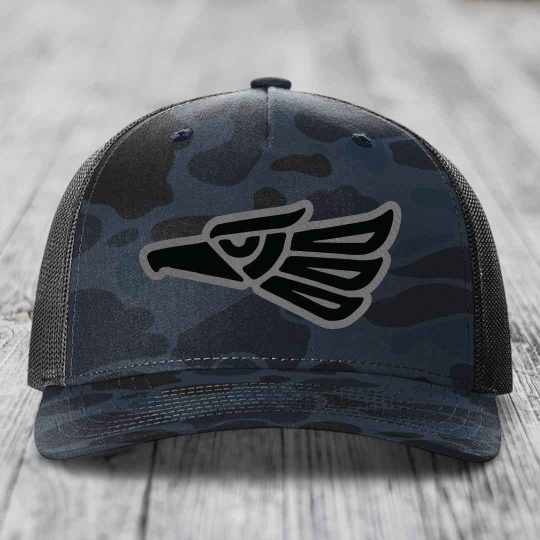 Camouflage trucker hat with black Aztec eagle patch on front.