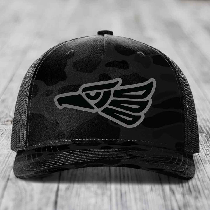 Black camouflage trucker hat with grey Aztec eagle patch on front.