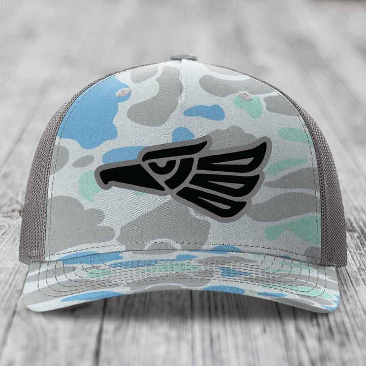 Camouflage trucker hat with black eagle patch on front, gray mesh back, resting on wooden surface.