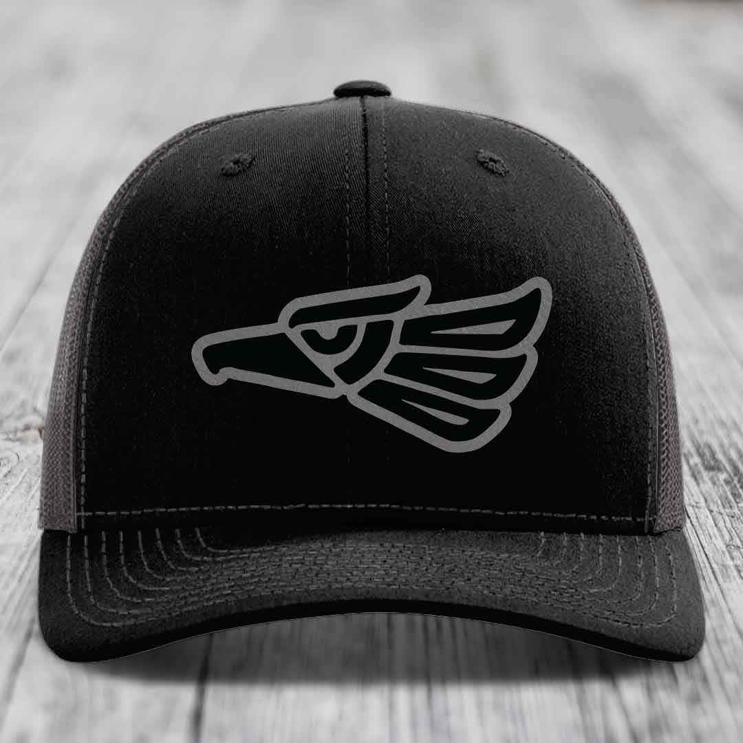 Black trucker hat with grey Aztec eagle patch on front, resting on wooden surface.