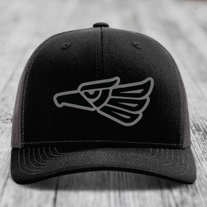 Black trucker hat with grey Aztec eagle patch on front, resting on wooden surface.