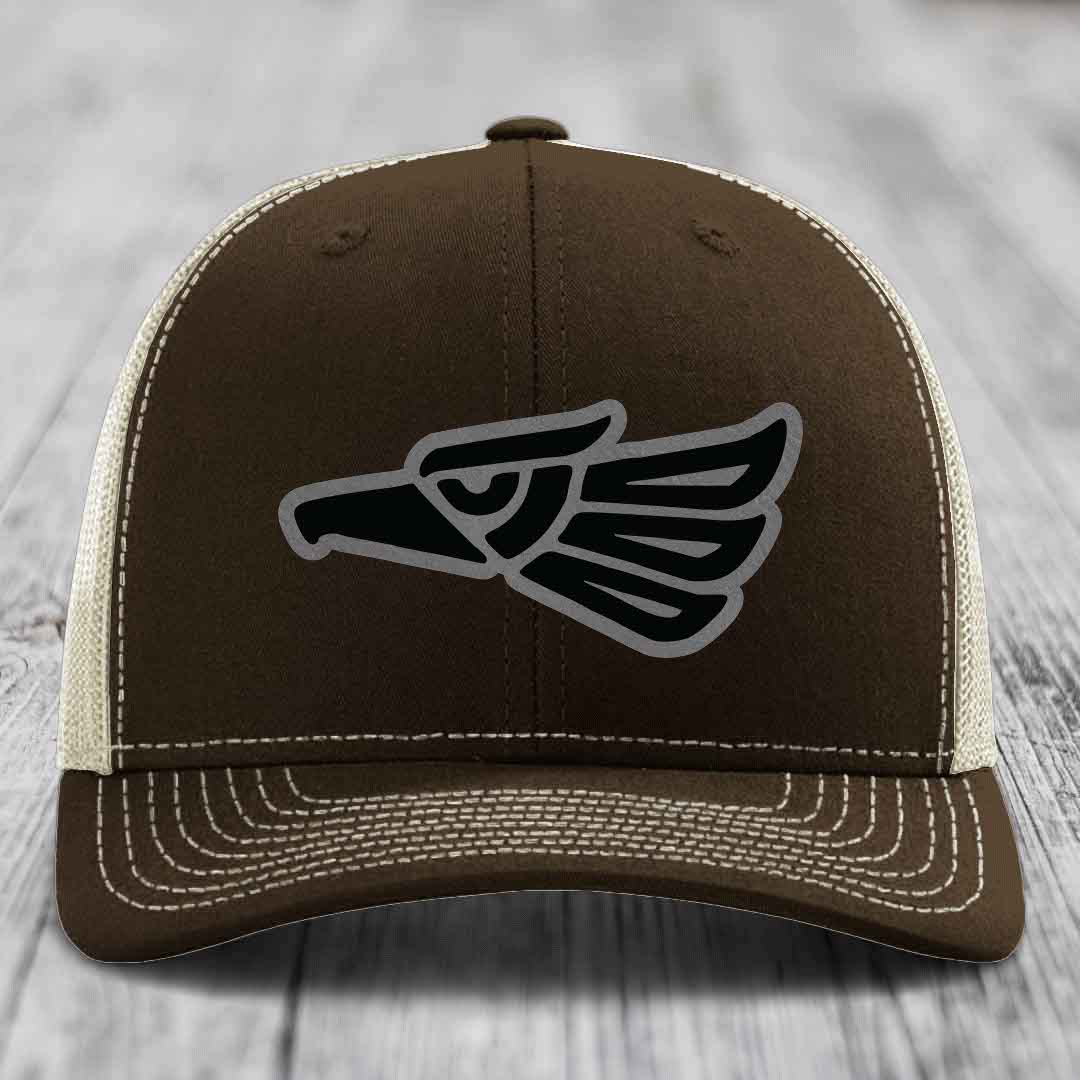 Brown trucker hat with black and grey eagle patch on front, white stitching, mesh back.