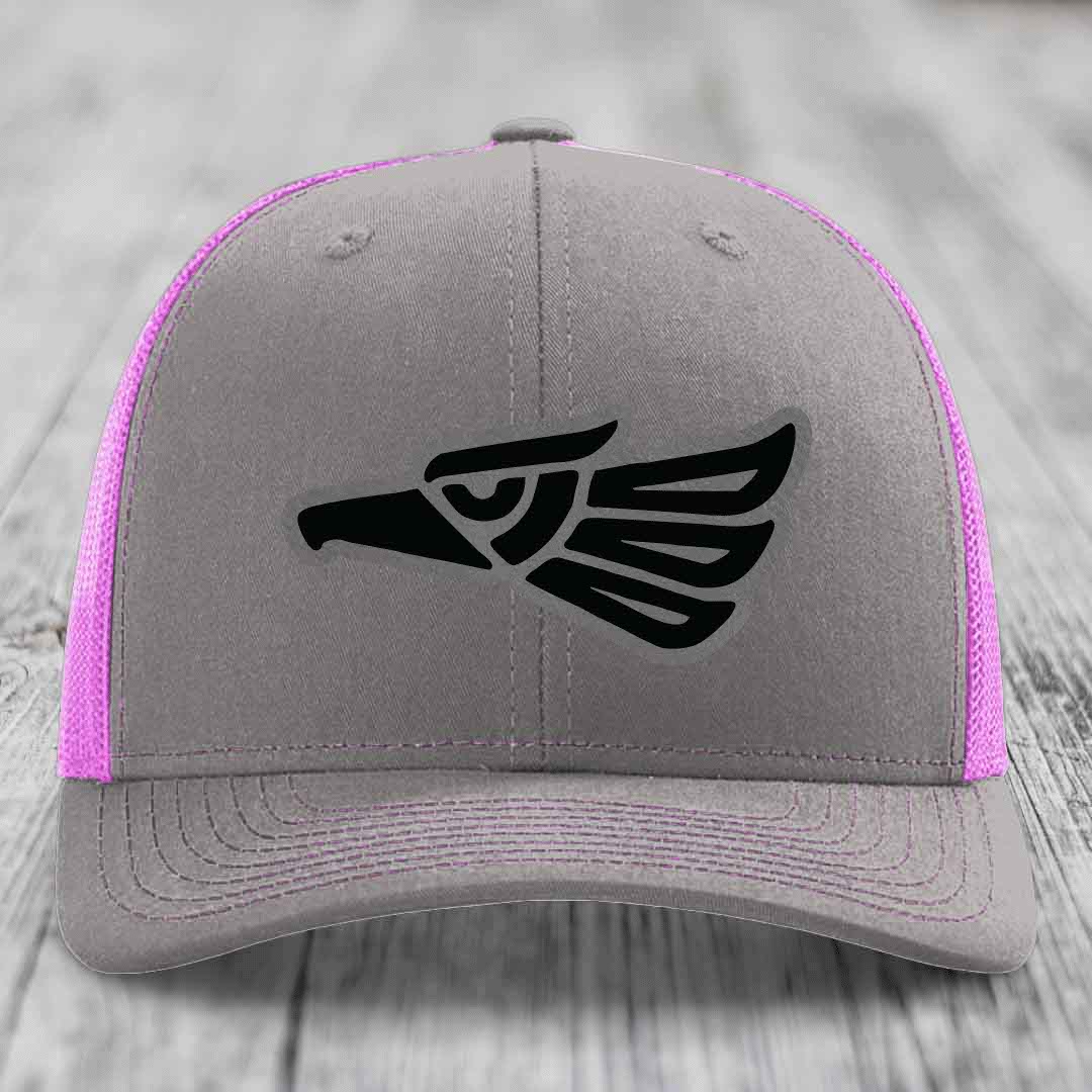 Grey trucker hat with black Aztec-style eagle patch and pink mesh sides.