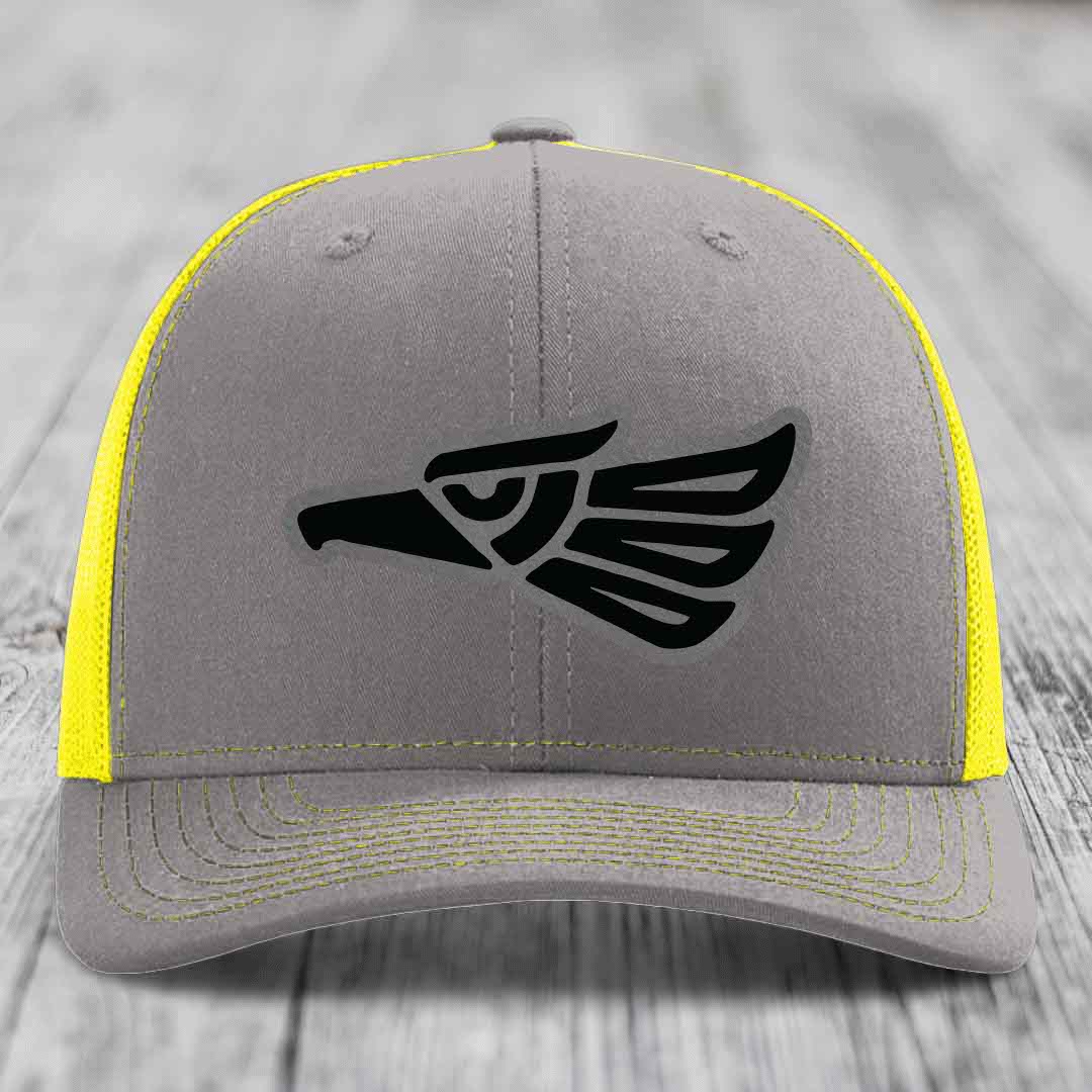Grey trucker hat with black Aztec eagle patch and yellow mesh back and stitching.