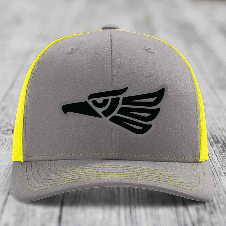 Grey trucker hat with black Aztec eagle patch and yellow mesh back and stitching.