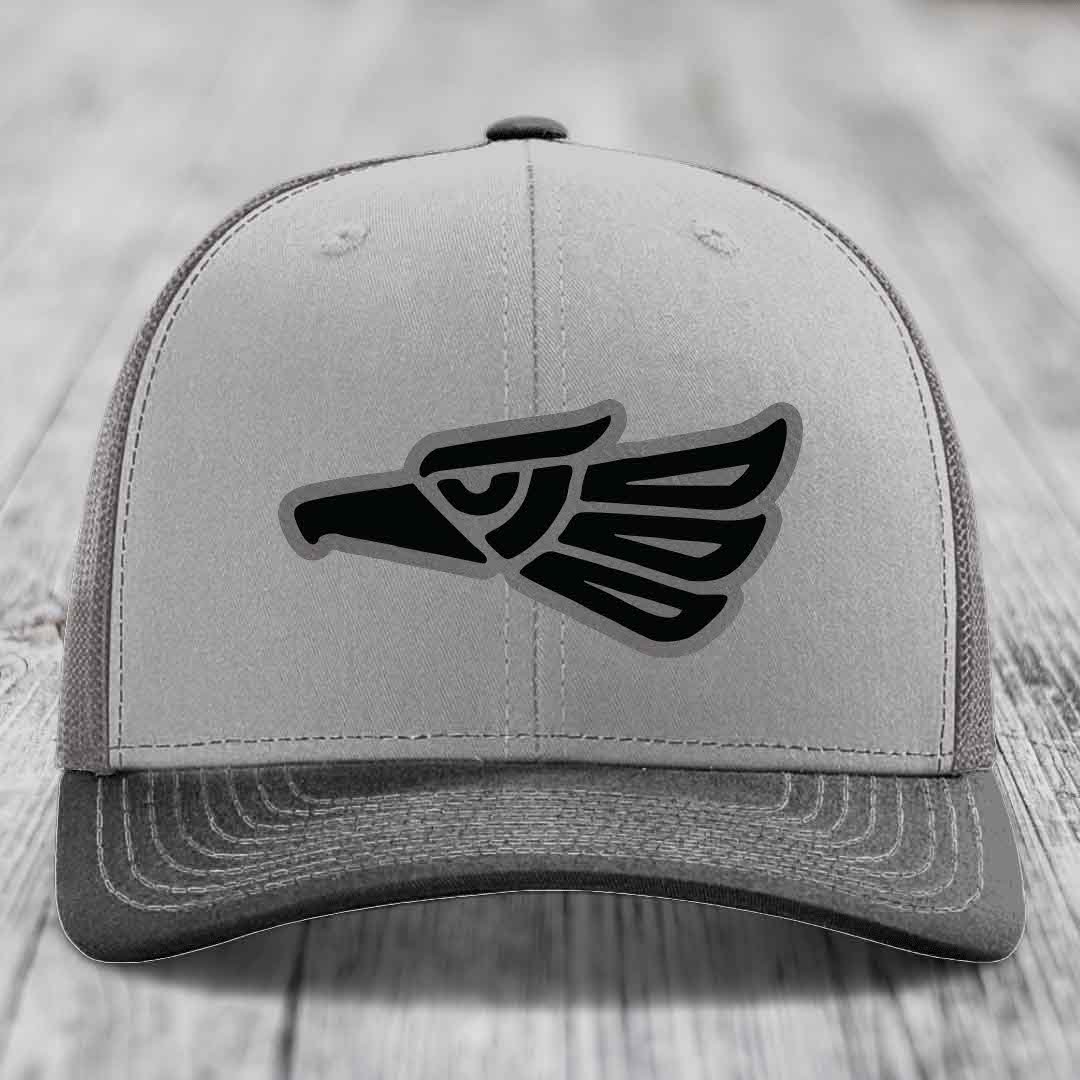 Grey trucker hat with black Aztec eagle patch on front, placed on wooden surface.