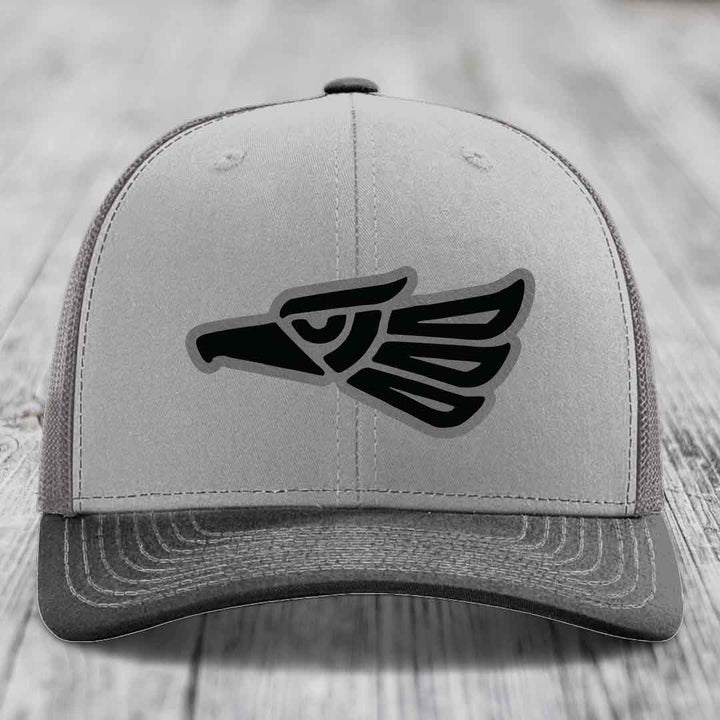 Grey trucker hat with black Aztec eagle patch on front, placed on wooden surface.