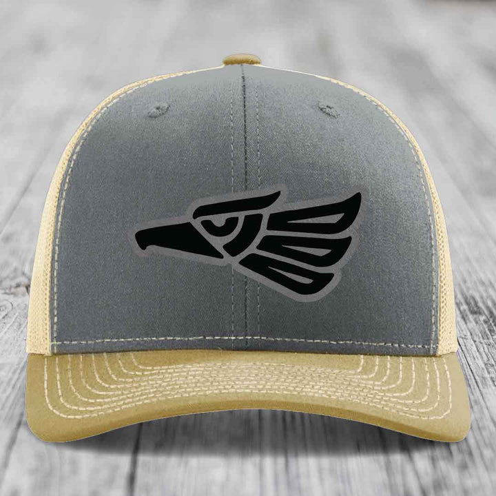 Grey and beige trucker hat with black Aztec eagle leather patch on front.