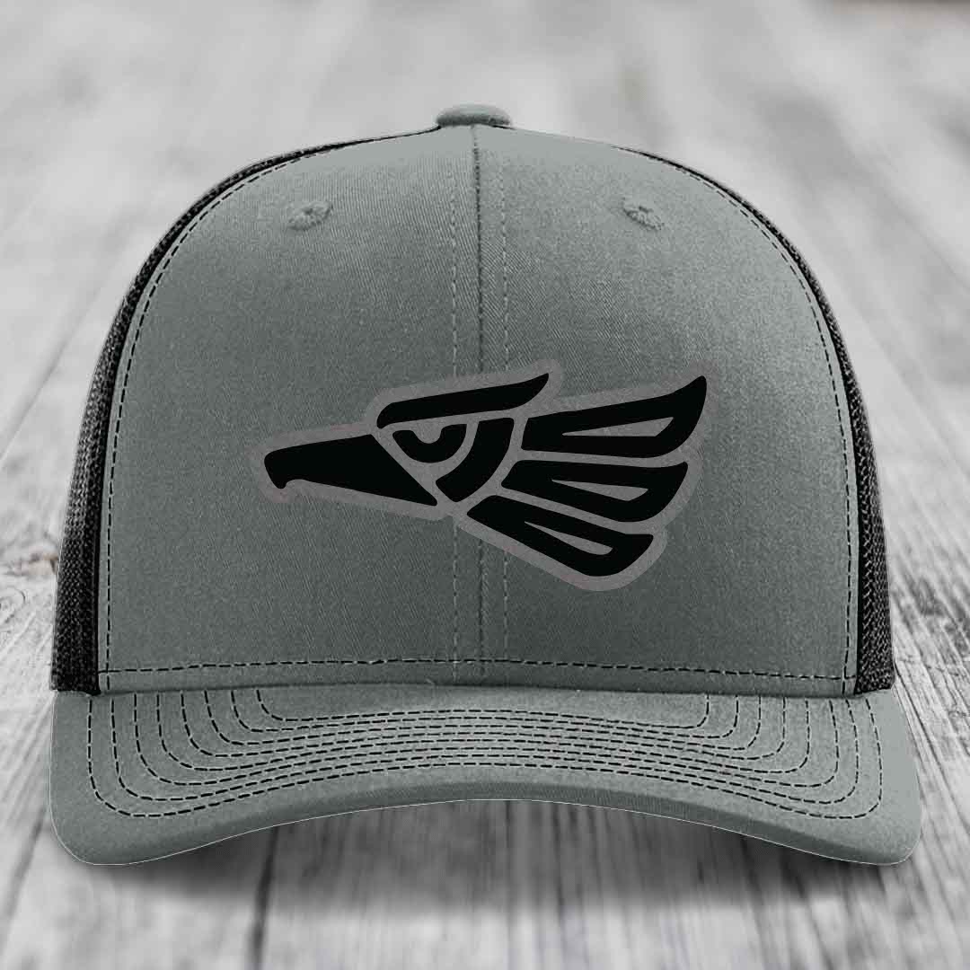 Grey trucker hat with black Aztec-style eagle patch on front, black mesh back, sitting on wooden surface.