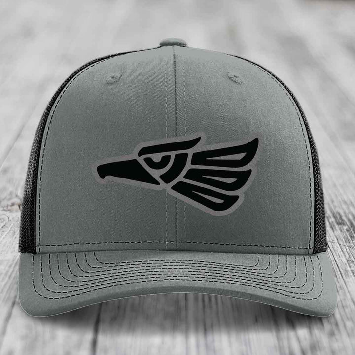 Grey trucker hat with black Aztec-style eagle patch on front, black mesh back, sitting on wooden surface.