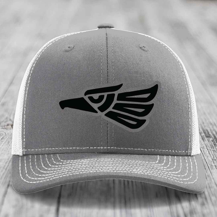 Grey trucker hat with black Aztec-style eagle patch on front, white mesh back and white stitching.