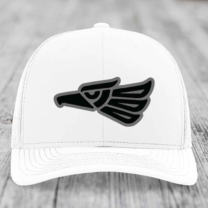 White trucker hat with black and grey Aztec eagle leather patch on front.